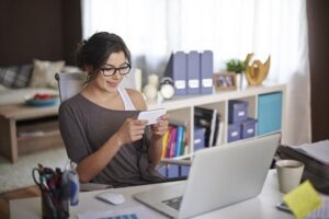 An image of a Virtual PA with glasses at home at her desk with her laptop, phone, pencils and coffee.