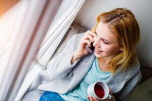 A close up on a woman in loungewear, holding a cup of tea and talking on a mobile phone while looking out a window.