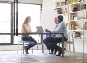 Three colleagues in business wear sitting around a table having a meeting in a glass-walled office space.
