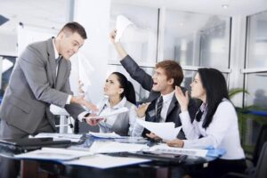 A man in an office with three of his colleagues sitting down at a desk while dealing with stress in the workplace.