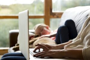 A woman using a laptop on a sofa at home with a child sleeping in the background, attempting to balance work from home.
