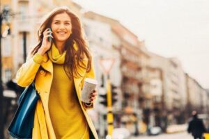 A PA in London in a yellow dress and coat smiling and talking on the phone while holding coffee and a handbag.