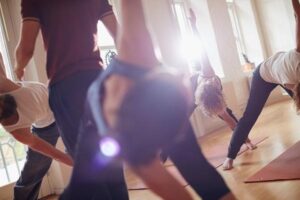 A group of people stretching in a yoga class with an instructor helping perfect the stretch.