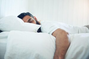 A man with dark hair and a beard in pyjamas sleeping on a white bed, bedsheet, duvet and pillow.