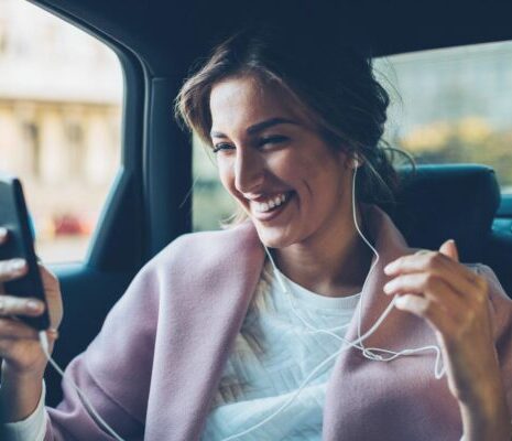 Young woman in a private role with a smartphone and headphones on the back seat of a car, smiling into her phone.