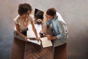 Two smiling colleagues talking in an informal setting with coffees and papers.