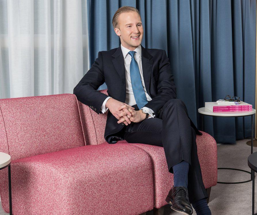 William Hanson, Etiquette Expert, sitting on a pink sofa with his legs crossed and arms folded in a classy manner.