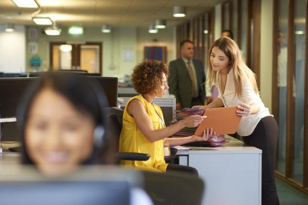 a young office junior takes a file from her supervisor for filing in a large open plan office . Co-workers can be seen defocussed in the foreground and background .