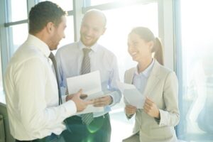 Three colleagues smiling together while in a meeting next to the windows of a well-lit office building.