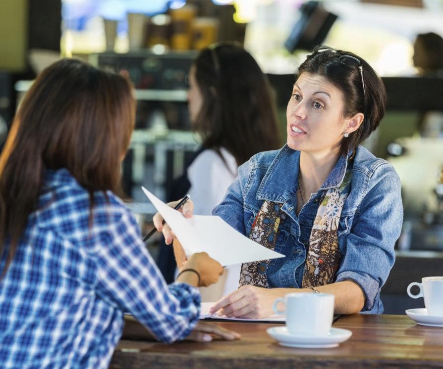 A woman conducting an appraisal of a private household staff member in a cafe, while wearing a denim jacket.
