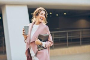 An elegant young temp wearing a pink coat walking on the sidewalk in an urban setting and looking back behind her shoulder.