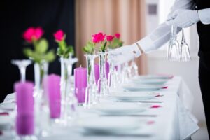 A butler places water glasses on a white tablecloth in a mansion, which is one of the qualities every butler should possess.