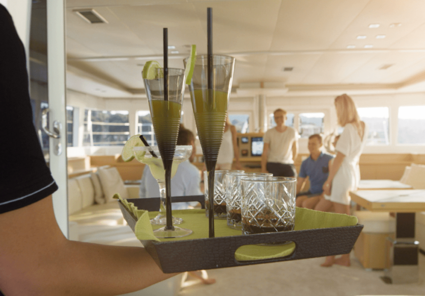A yacht worker carrying a tray of cocktails and champagne glasses over to a family vacationing on the yacht.