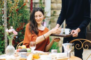 A young woman in an orange top sitting at a table outside being served by a modern-day butler.