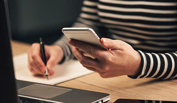 Woman holding mobile and writing down something with a laptop in front of her