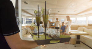 A temporary worker carrying a tray of drinks on a yacht towards a principal and their family.