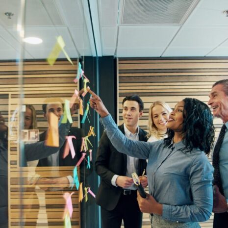 A female operations manager is standing with a group of work colleagues while she works on post-it notes on a glass window.