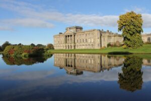 A large manor house where live-in couples work - there is a lake in the foreground and a few large trees surrounding it.