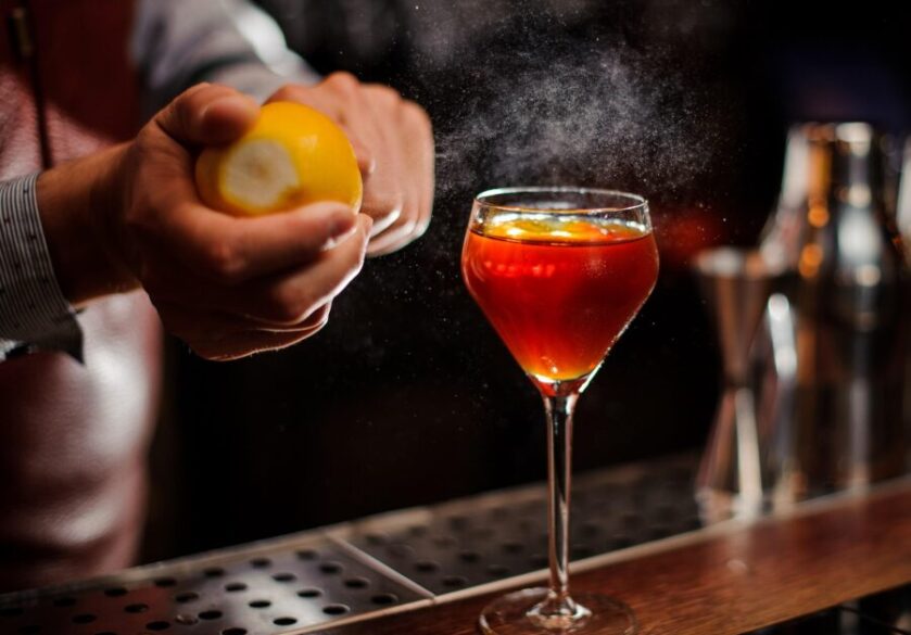 A bartender is adding lemon zest to the red cocktail at a modern bar's counter. He carefully crafts the drinks.