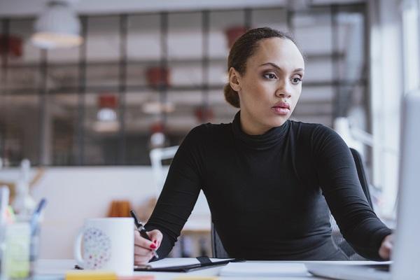 Image of african young woman working new business assignment. Female executive sitting at her desk using laptop and writing notes at office.
