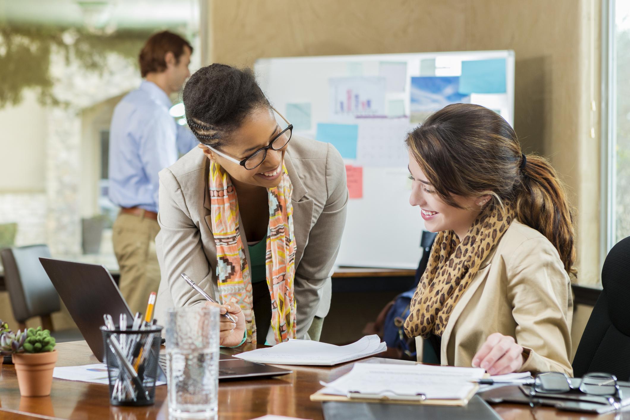 Cheerful mid adult African American and young Caucasian businesswomen discuss something in the office. They are reviewing documents at the Caucasian woman's desk. A man is working in the background.