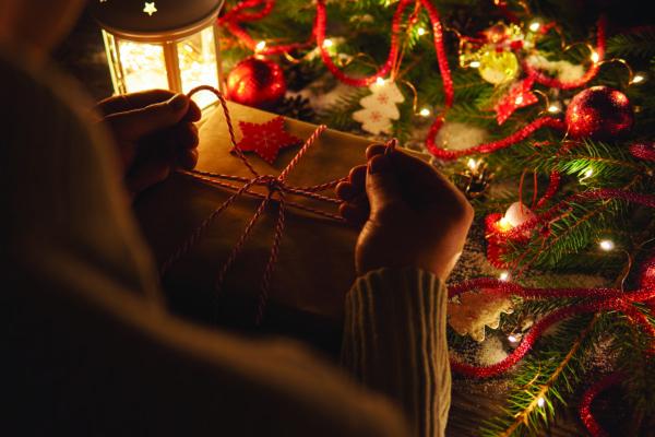 Crop shot of person tying up colorful twine on wrapped Christmas present at table with glowing garland