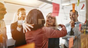 Two HR businesswomen are hugging and smiling while brainstorming with their colleagues in a bright, contemporary office space.