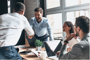 Four business professionals in a meeting room with two people stood, shaking hands