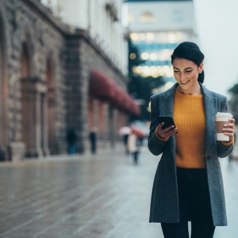 A woman walking on a city street holding a coffee and smiling at her phone.
