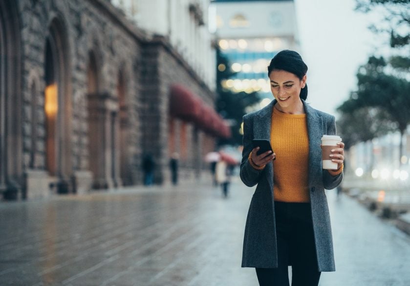A woman walking on a city street holding a coffee and smiling at her phone.
