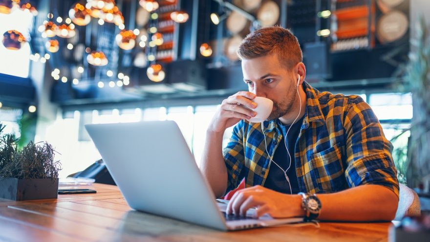 Young Caucasian blogger with earphones in ears and in plaid shirt drinking coffee and using laptop while sitting in coffee shop.