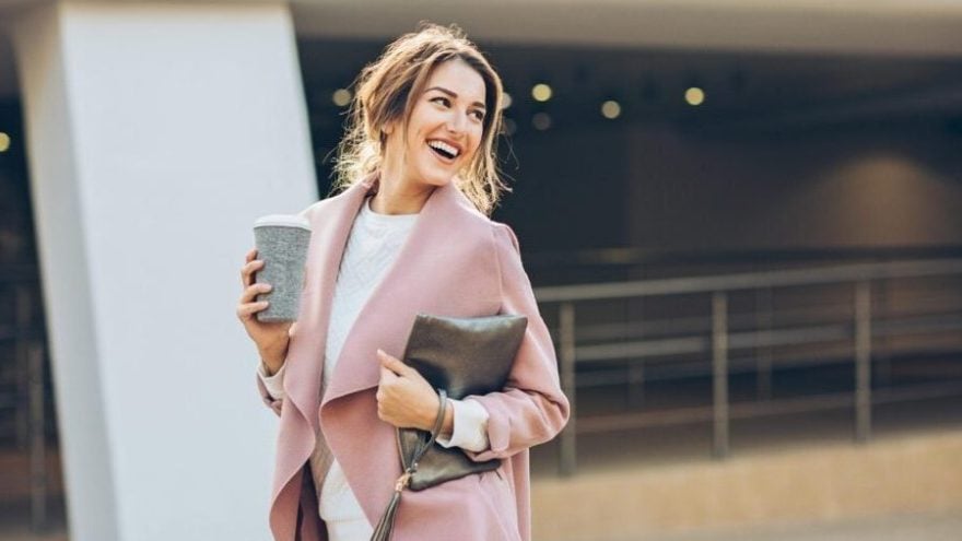 A woman in a pink coat walks outside with a coffee
