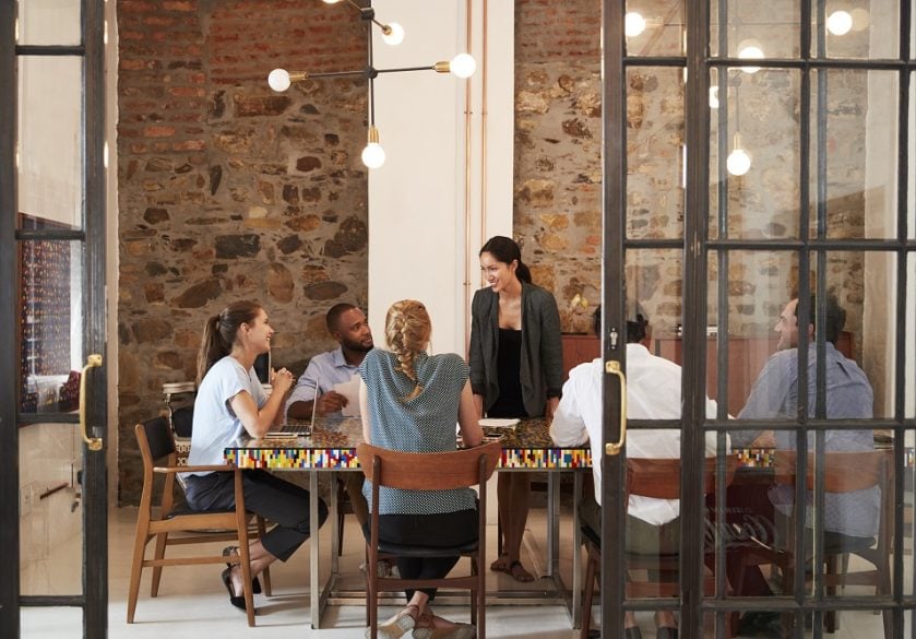A human resources team of five people in a creative office with exposed brick wall and hanging lights having a group meeting