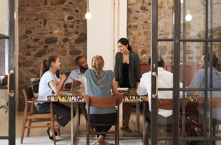 A human resources team of five people in a creative office with exposed brick wall and hanging lights having a group meeting