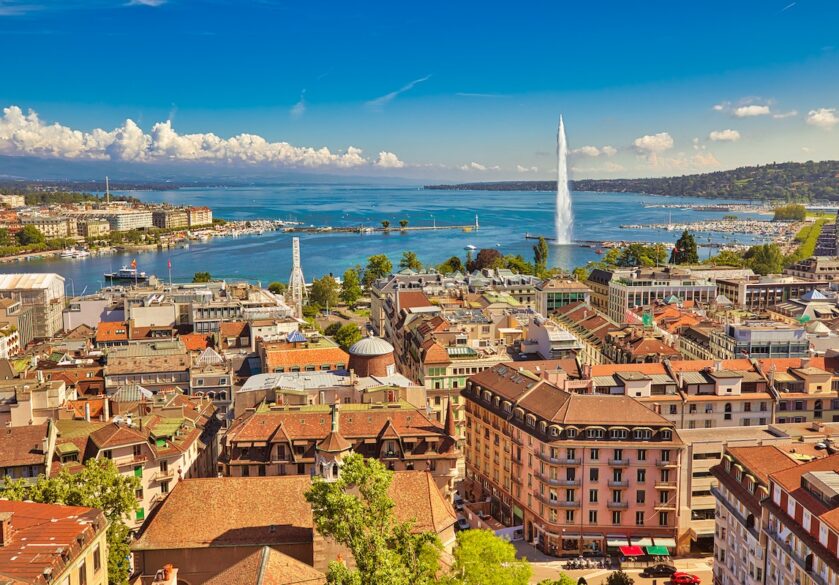 old buildings in front of a stretch of water with a fountain coming out of it - the skyline of Geneva.