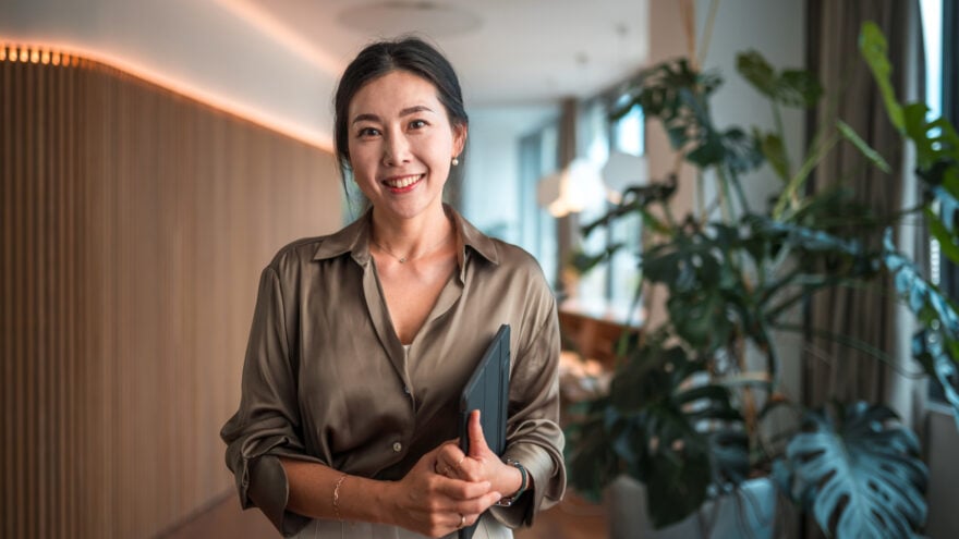 Positioned within a modern corporate environment, a mid-aged Asian businesswoman of striking beauty stands in her office, poised with a digital tablet, using it to execute digital tools and wireless technology to perfection in her strategic endeavors.