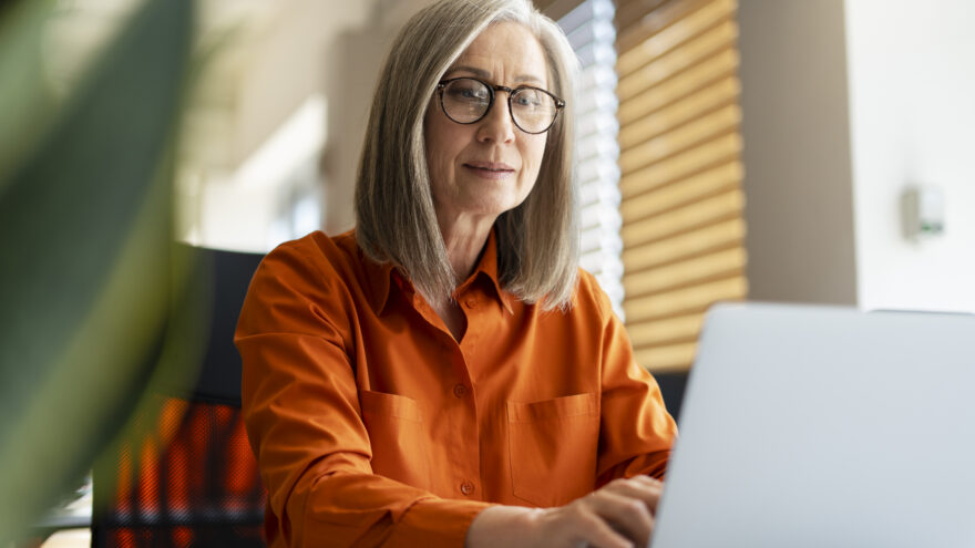 Portrait of confident serious mature businesswoman wearing stylish eyeglasses, orange shirt using laptop working online in modern office. Successful manager planning project, typing on keyboard